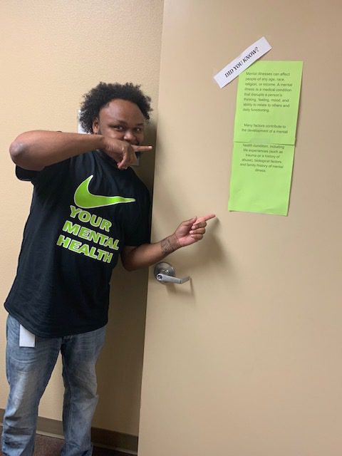 Quinta Anderson, counselor at the New Season Biloxi Treatment Center, designed the shirts to celebrate mental health awareness in May. Here he stands with one of the center’s informational posters to educate patients on the importance of mental health.
